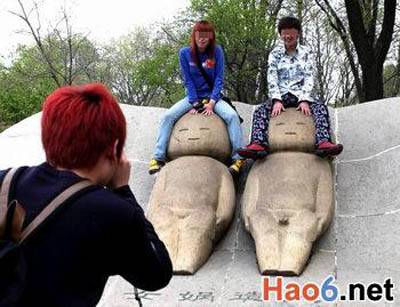 Two tourists sit on top of two sculptures to pose for a photo